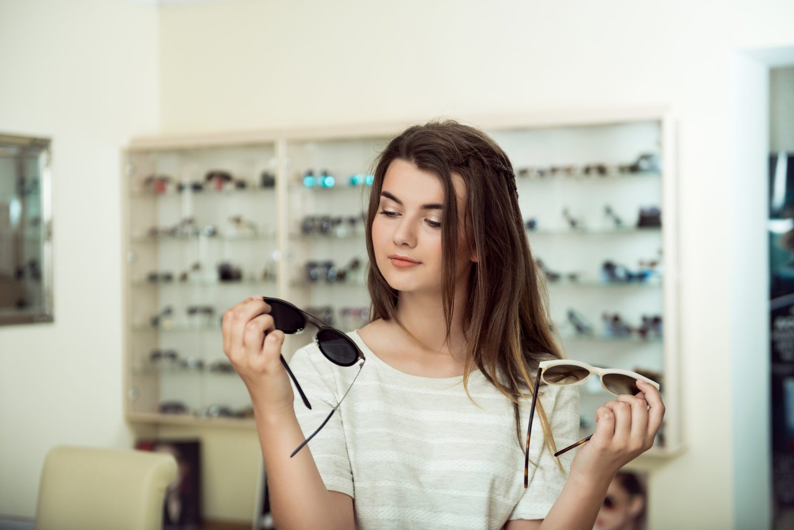 Which one is better fit me. Portrait of cheerful hesitating woman in optician store, making decision, holding stylish sunglasses, choosing what she should buy. Great discounts for buying two pairs.